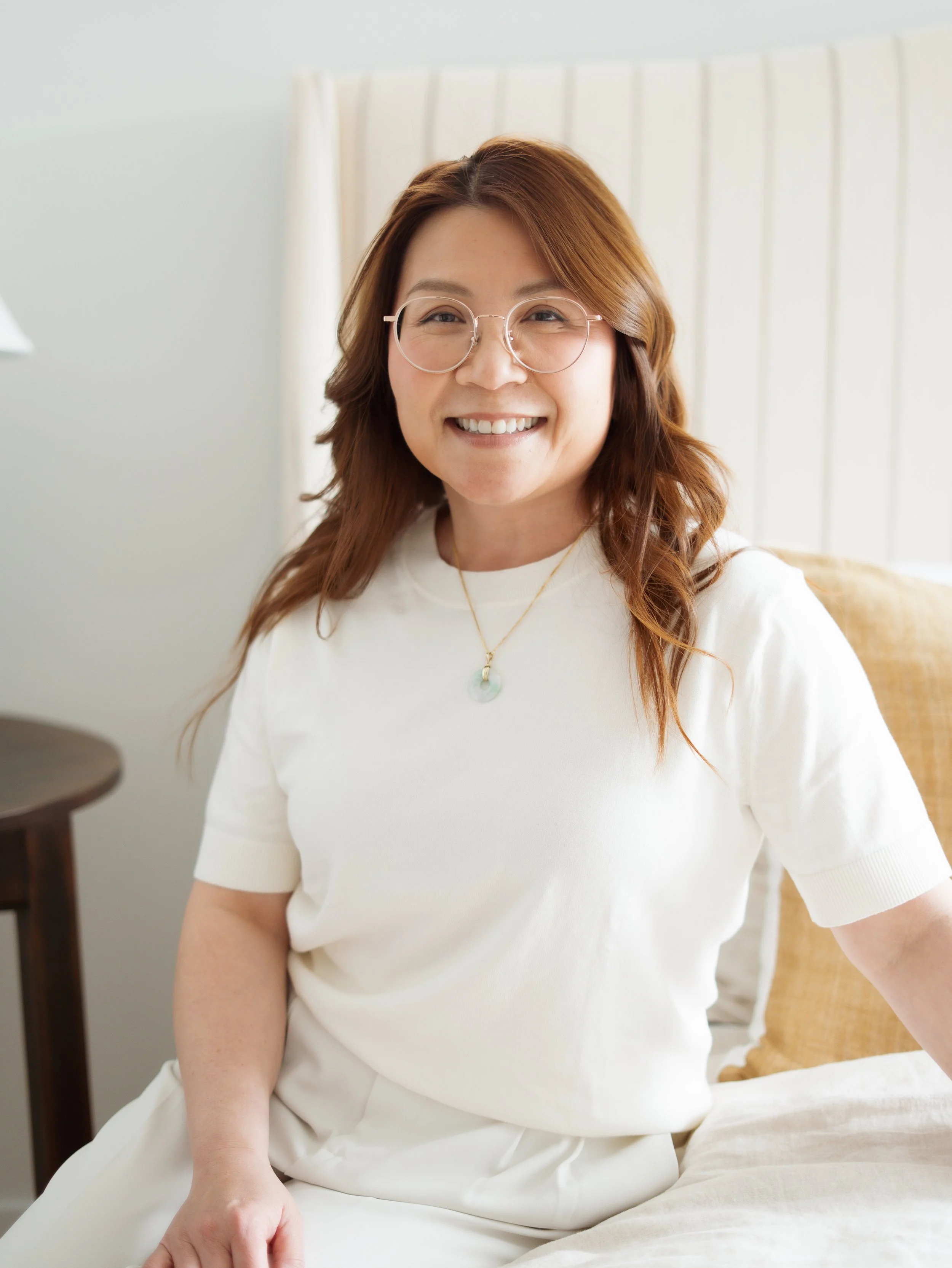A woman with brown wavy hair, wearing glasses and a white shirt, smiling while sitting on a bed in a well-lit room.