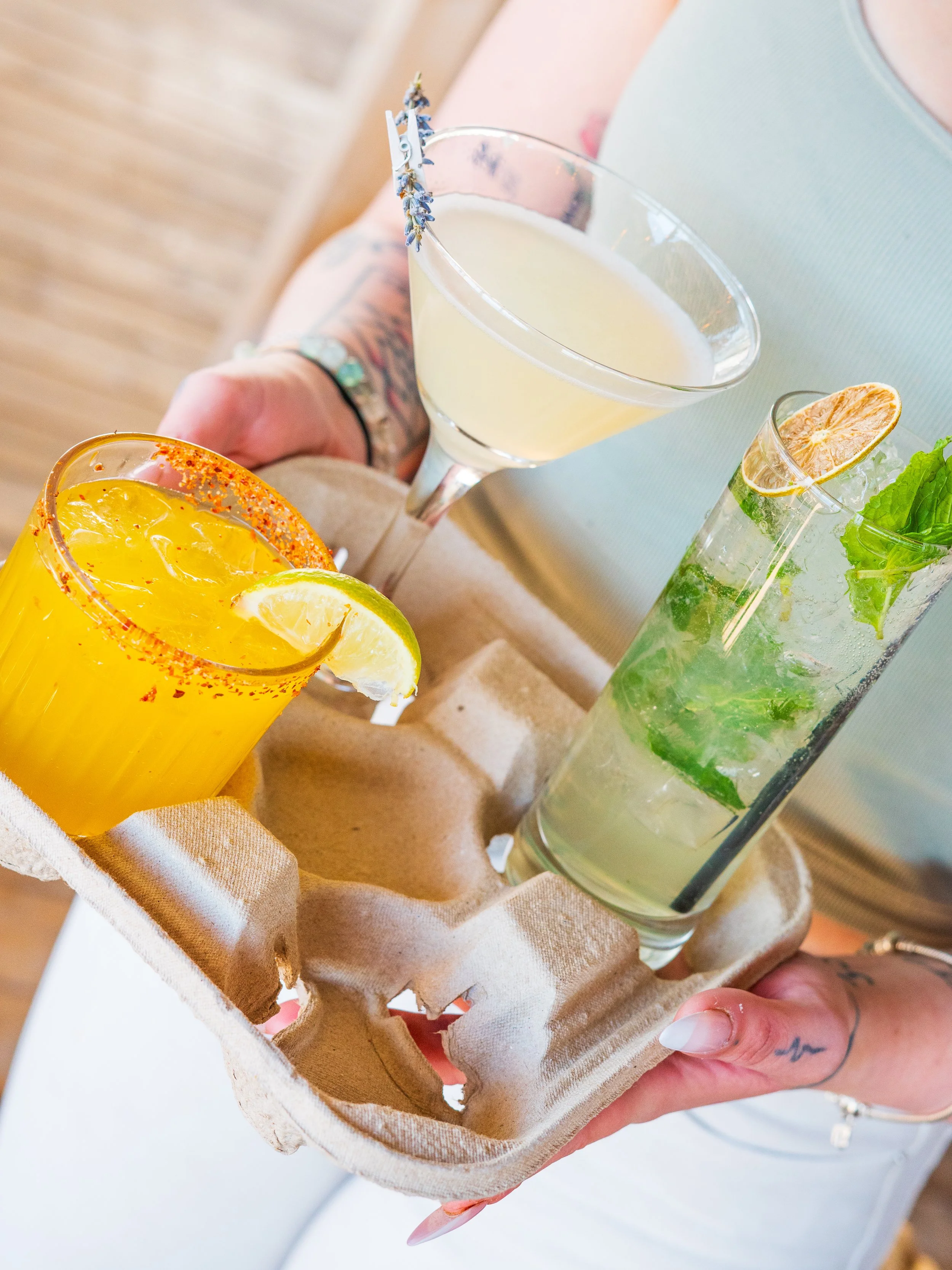 Person holding a tray with three different cocktails: a yellow drink with lemon slice and chili salt rim, a white or clear drink with lavender garnish, and a clear drink with lime, lemon, and mint leaves.