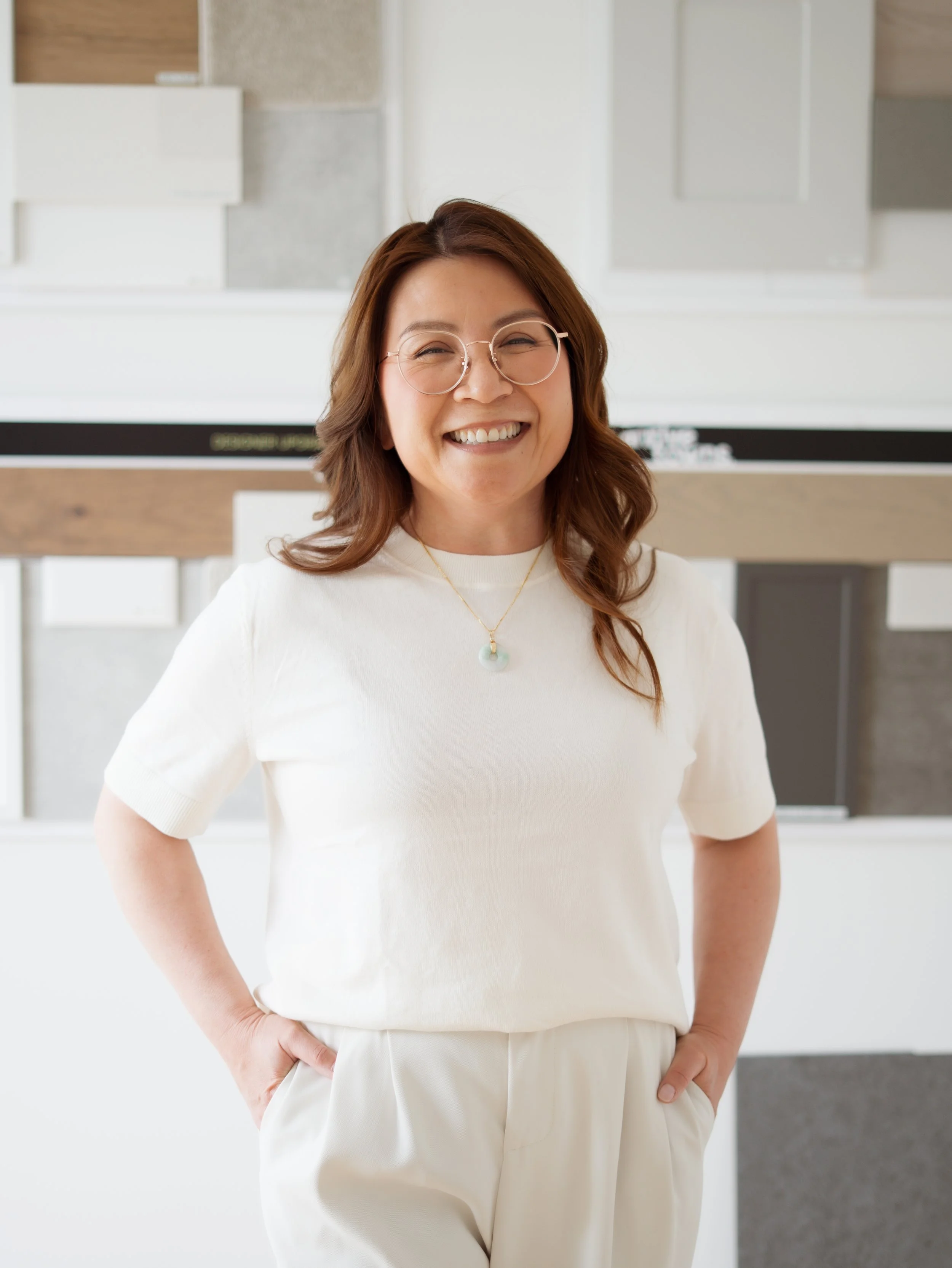 A woman with brown hair, round glasses, and a white top standing in a bright, modern space with her right hand on her hip, smiling at the camera.