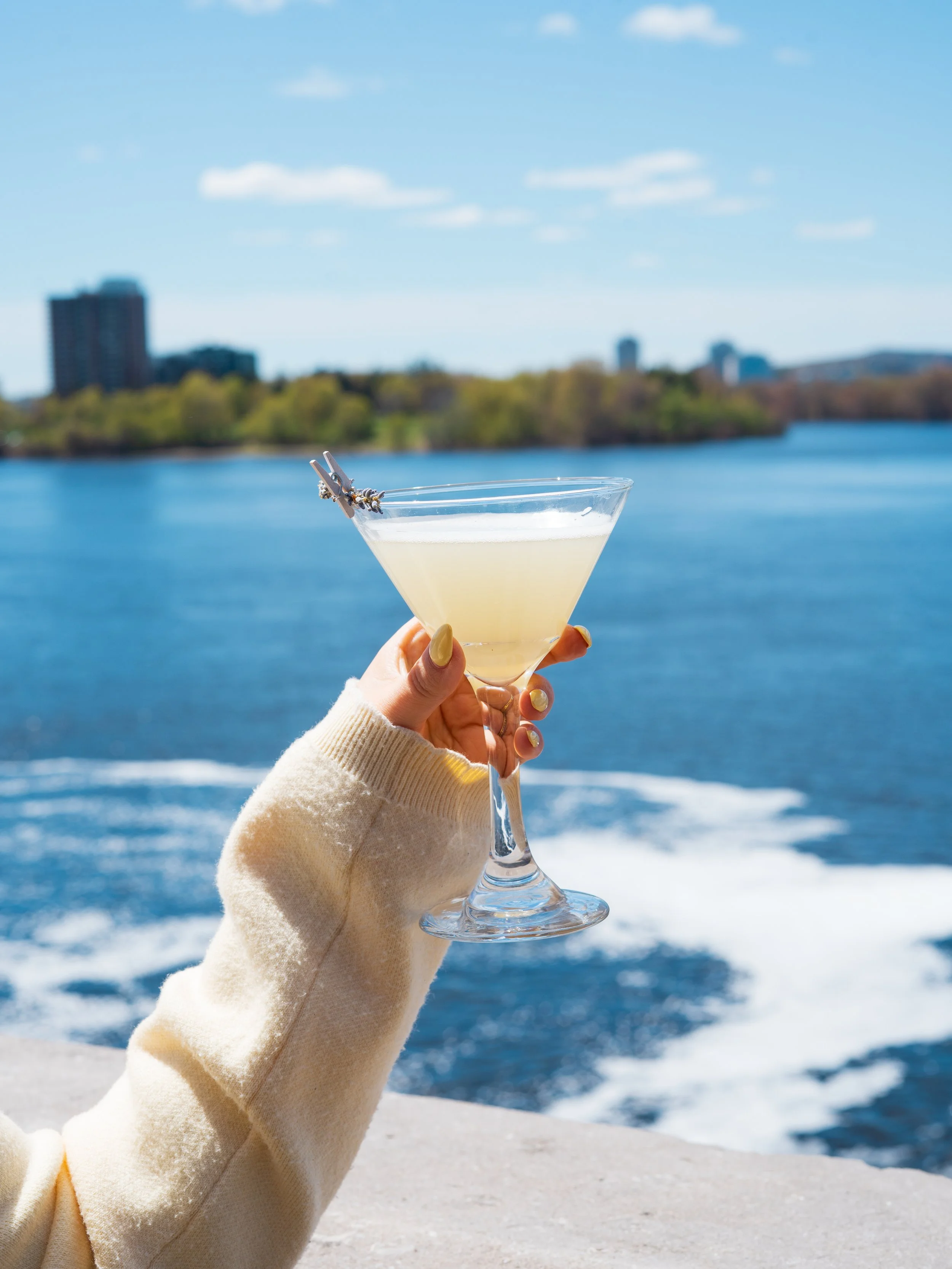 A person's hand holding a cocktail glass with a yellow drink and a small clothespin on the rim, with a lake, trees, and buildings in the background on a sunny day.