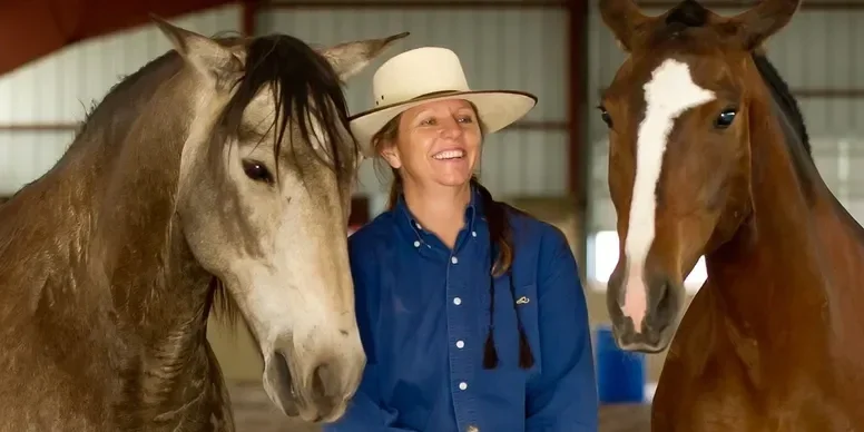 A woman in a blue shirt and wide-brimmed hat smiling between two horses inside a barn.