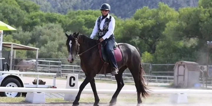 A person riding a horse on a dirt path at an outdoor equestrian facility, wearing a helmet and riding gear with trees in the background.