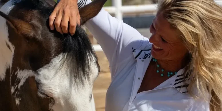 A woman with blonde hair and a turquoise necklace smiling while petting a dark brown and white horse.