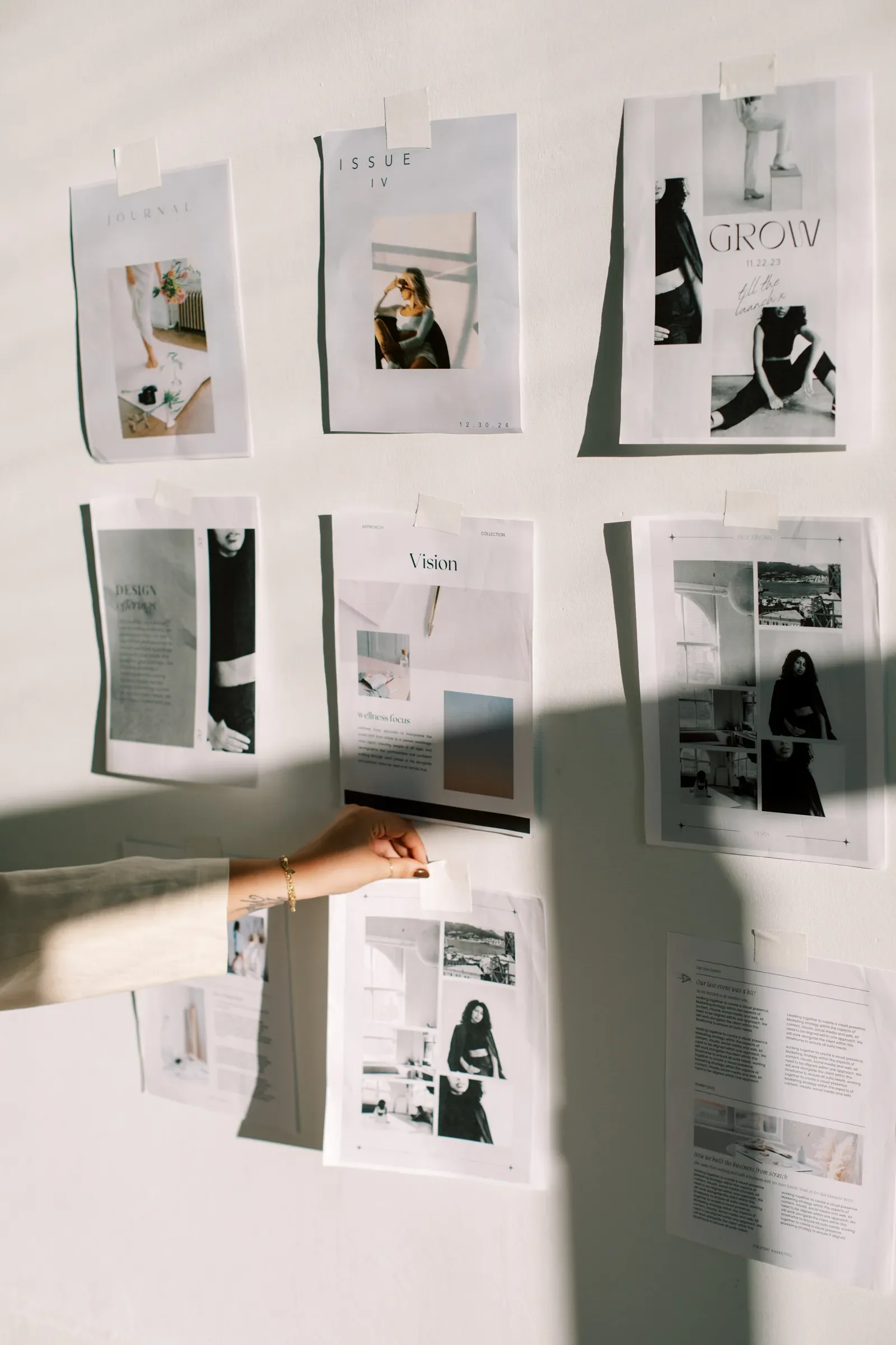 Display of multiple black and white photo prints and articles taped to a white wall, with a person holding one of the papers.