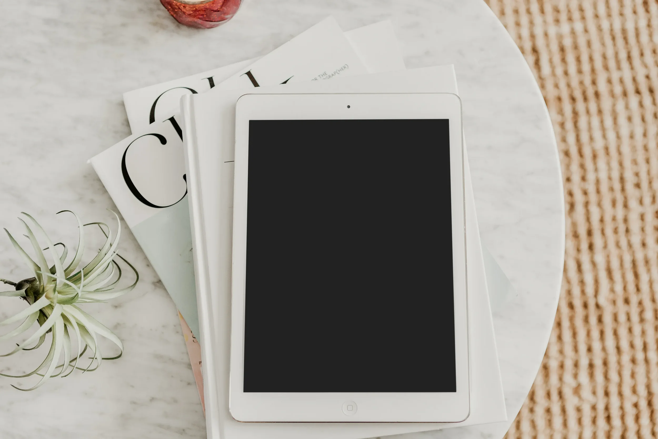A white tablet with a blank black screen placed on top of a white book, surrounded by a small plant, a magazine, and a white paper on a white marble surface. A glass container with red contents is in the upper left corner. A textured brown surface is partially visible on the right side.