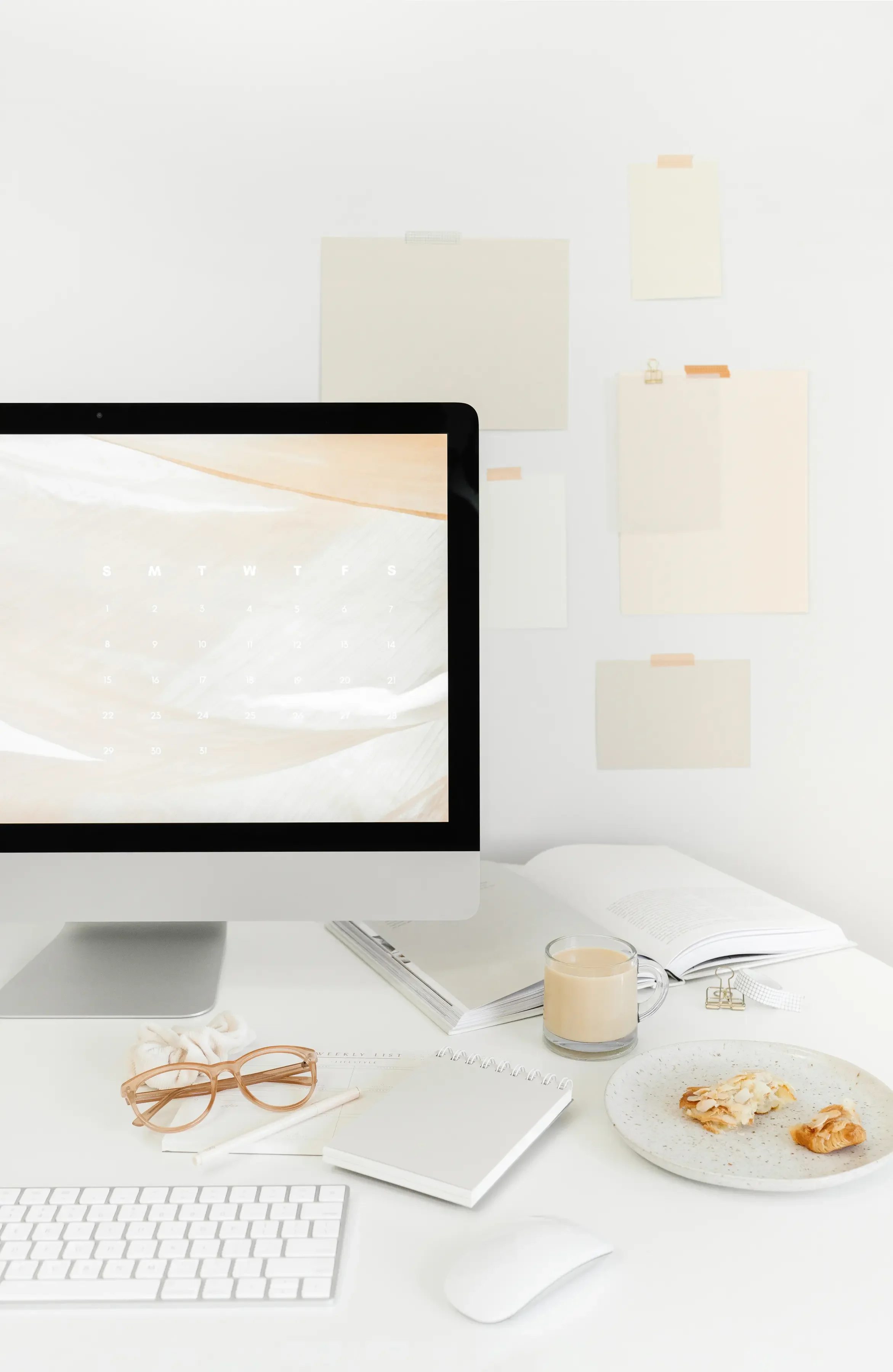 A minimalistic, white office desk setup with an iMac computer, a wireless keyboard, a mouse, a pair of pink glasses, an open notebook, a closed notepad, and a small plate with croissants. There is also a glass of coffee, an open book, and some papers with clips, against a white wall with beige sticky notes.