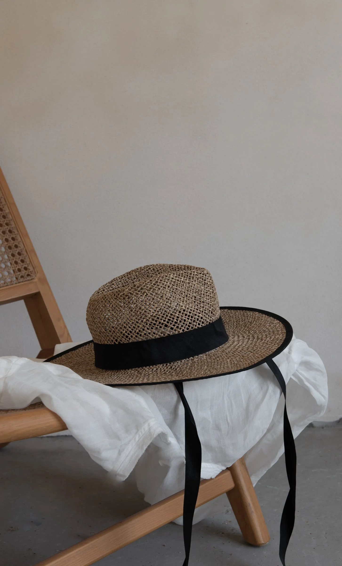A straw sun hat with a black band resting on a white bedding on a wooden bed frame. A portion of a wooden chair and a white wall are visible in the background.