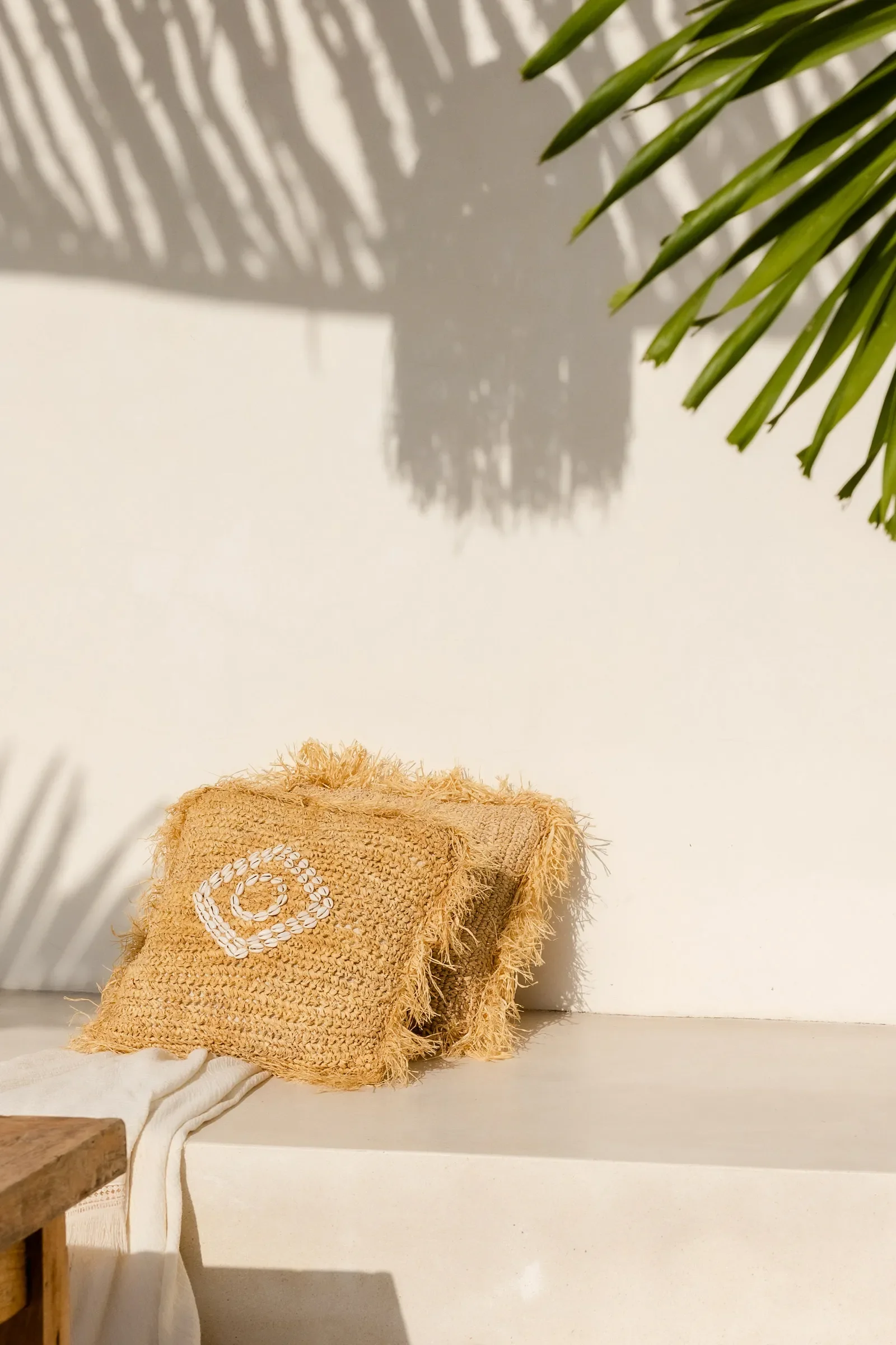A woven straw bag with a white circular pattern resting on a white surface, with a white cloth draped partially over the edge. Green palm leaves and their shadows are visible in the background, and a beige fluffy object is at the bottom of the bag.
