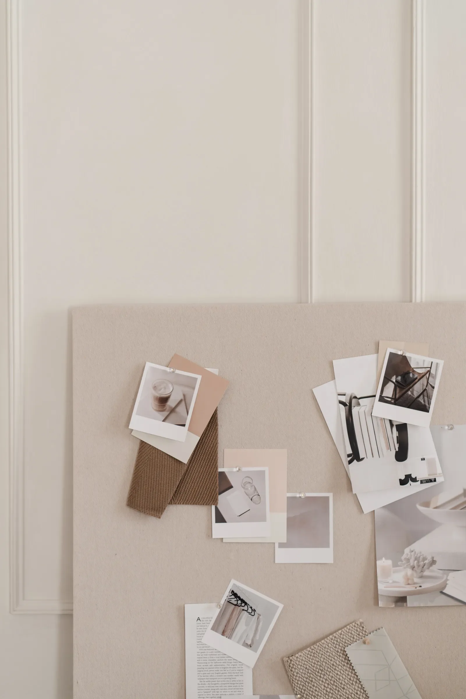 A beige-colored board with various photographs, fabric swatches, and paper pinned or taped to it, with a neutral wall and white side table in the background.