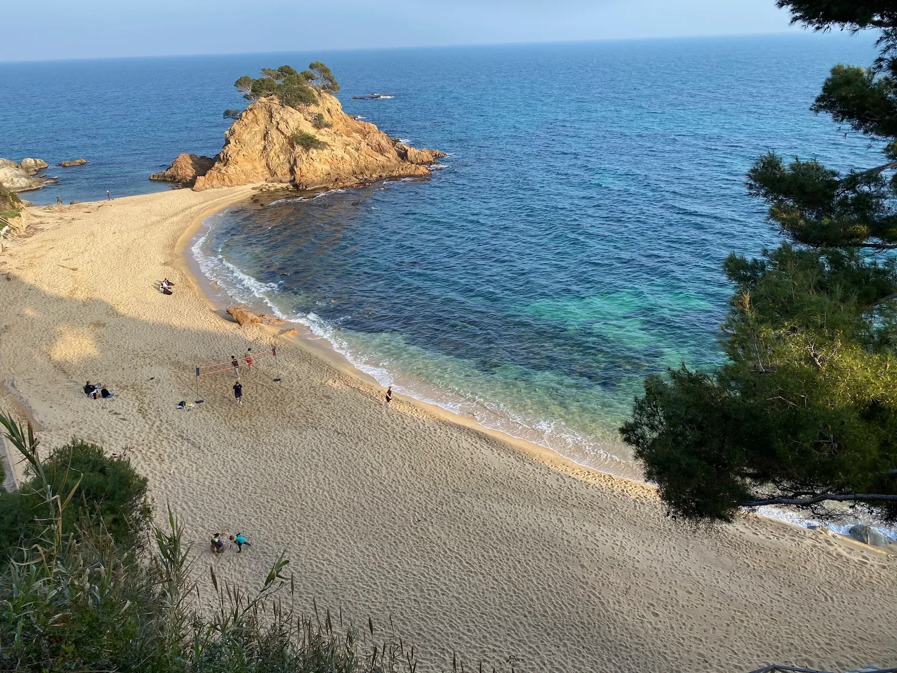 A sandy beach with a large rock formation and trees, overlooking the ocean with clear water near the shore, and people walking and playing on the beach.