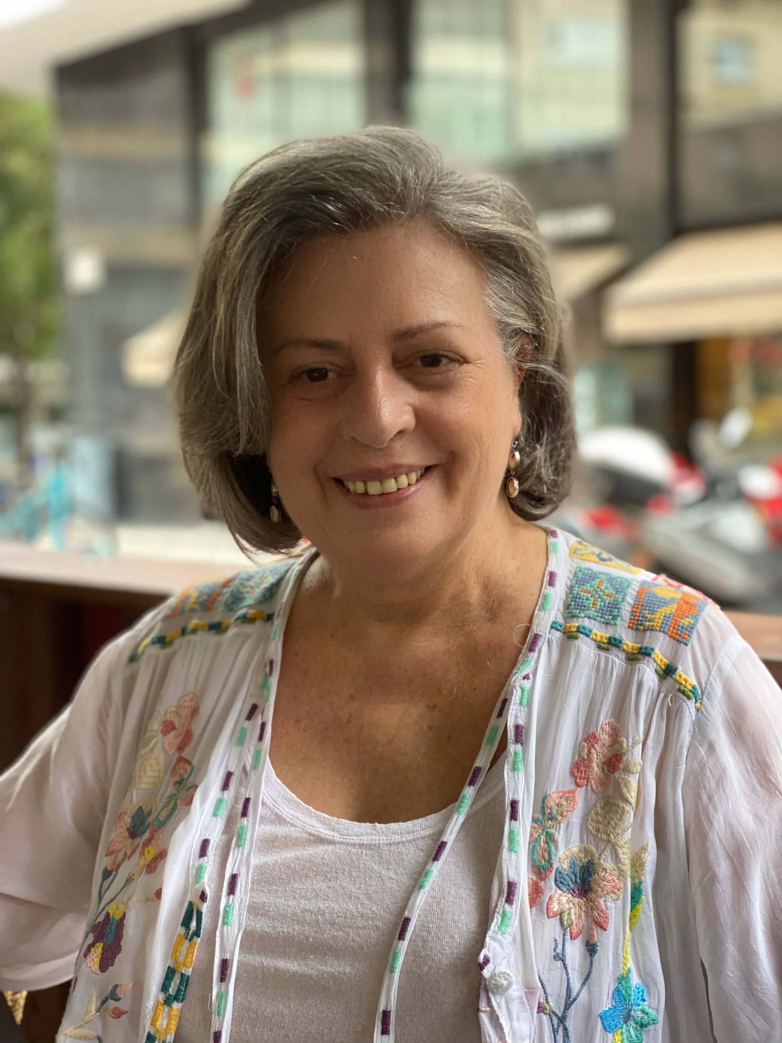 A smiling woman with short gray hair, wearing a white embroidered top, sitting inside a restaurant or cafe with large windows and a street view in the background.