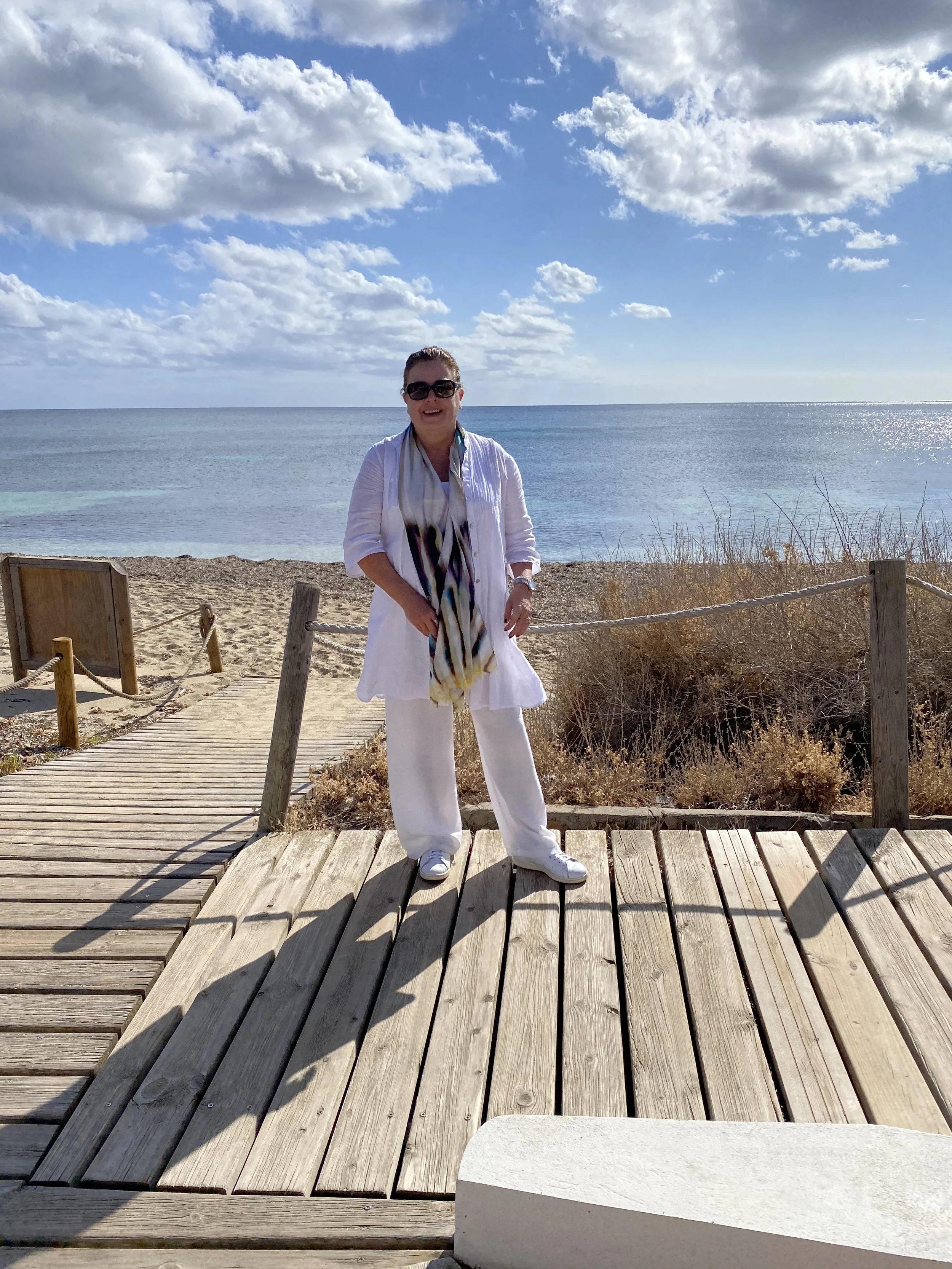 A woman in white clothing, sunglasses, and a scarf standing on a wooden pathway at the beach with the ocean and cloudy sky in the background.