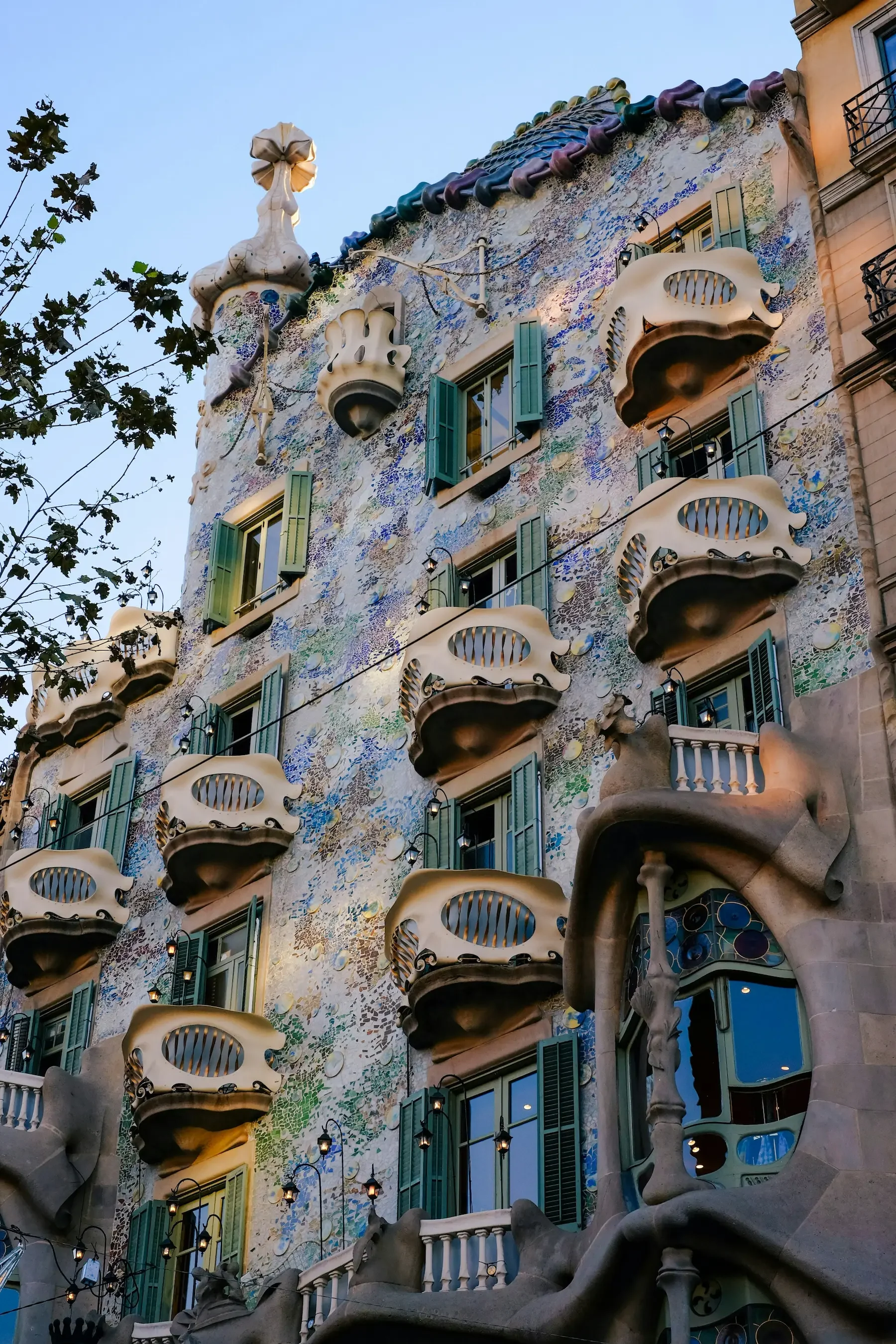 The facade of Casa Batlló, a famous building in Barcelona designed by Antoni Gaudí, features colorful mosaic tiles, unique bone-like balconies, and ornate rooftop sculptures resembling a dragon or sea creature.