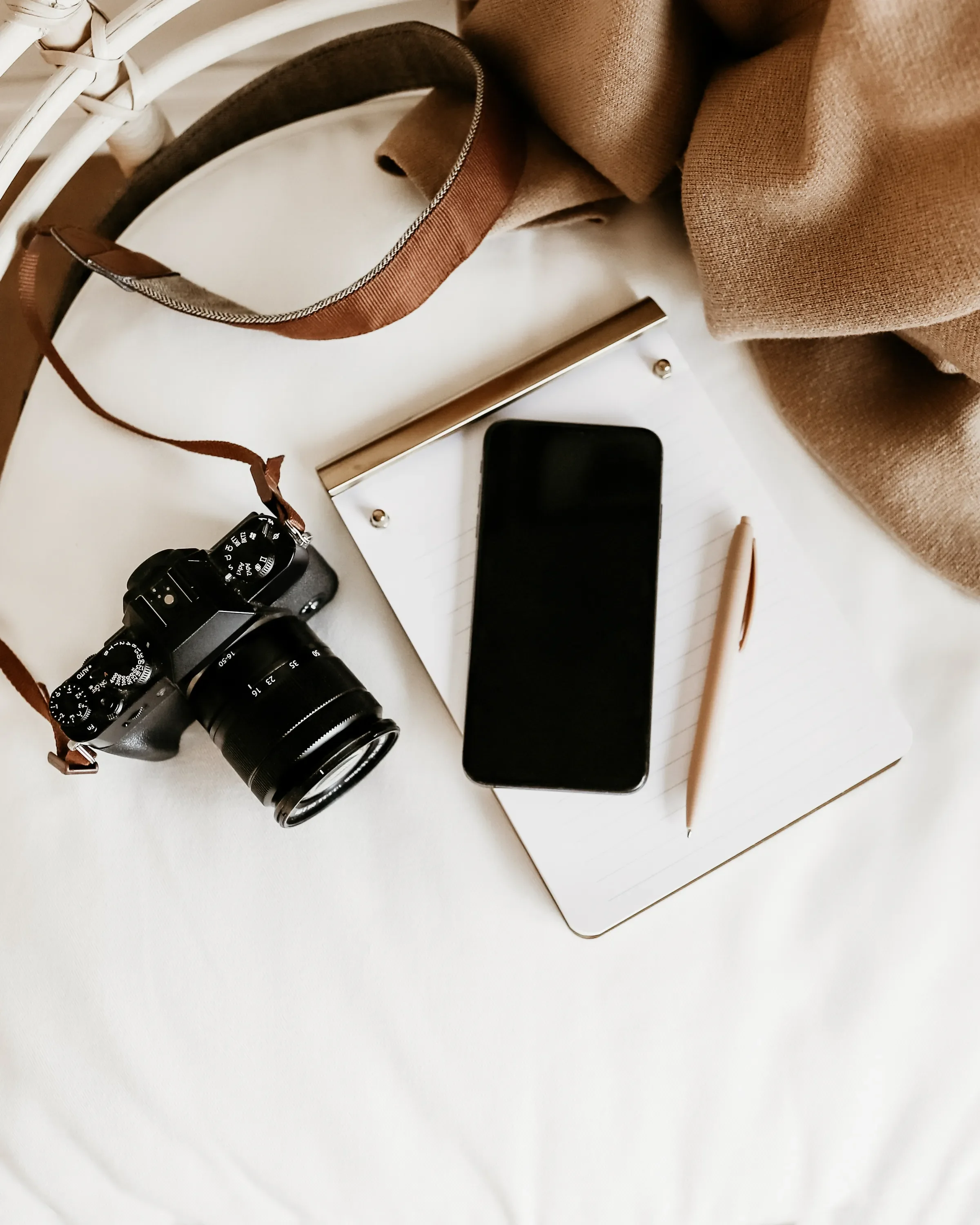 Array of objects on a white table including a camera, smartphone, notebook with a pen, a beige scarf, a brown strap, keys, and a window frame.