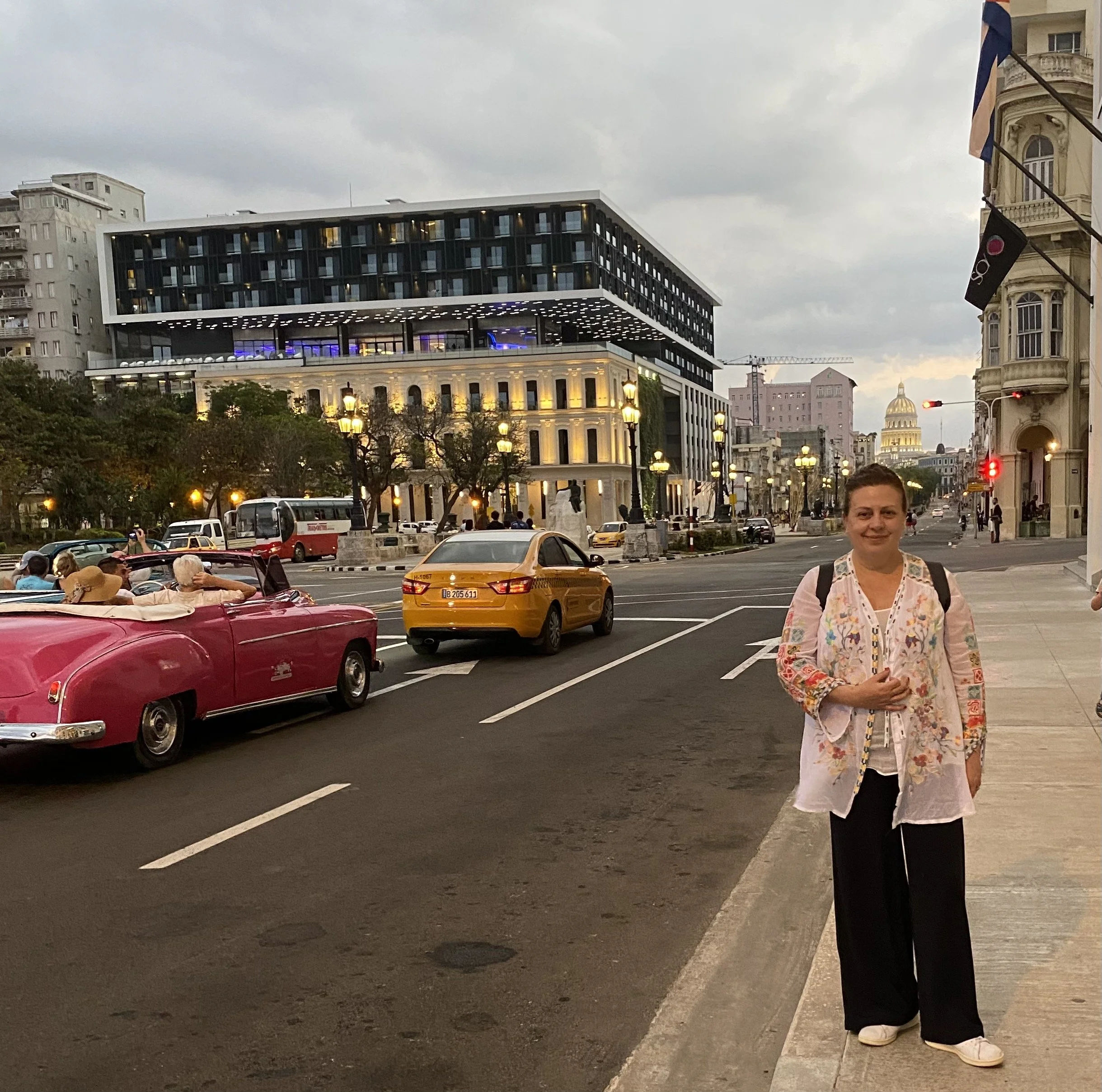 A city street scene with classic and modern buildings, vintage and modern cars, street lamps, and a woman wearing a floral jacket and black pants standing at the curb.