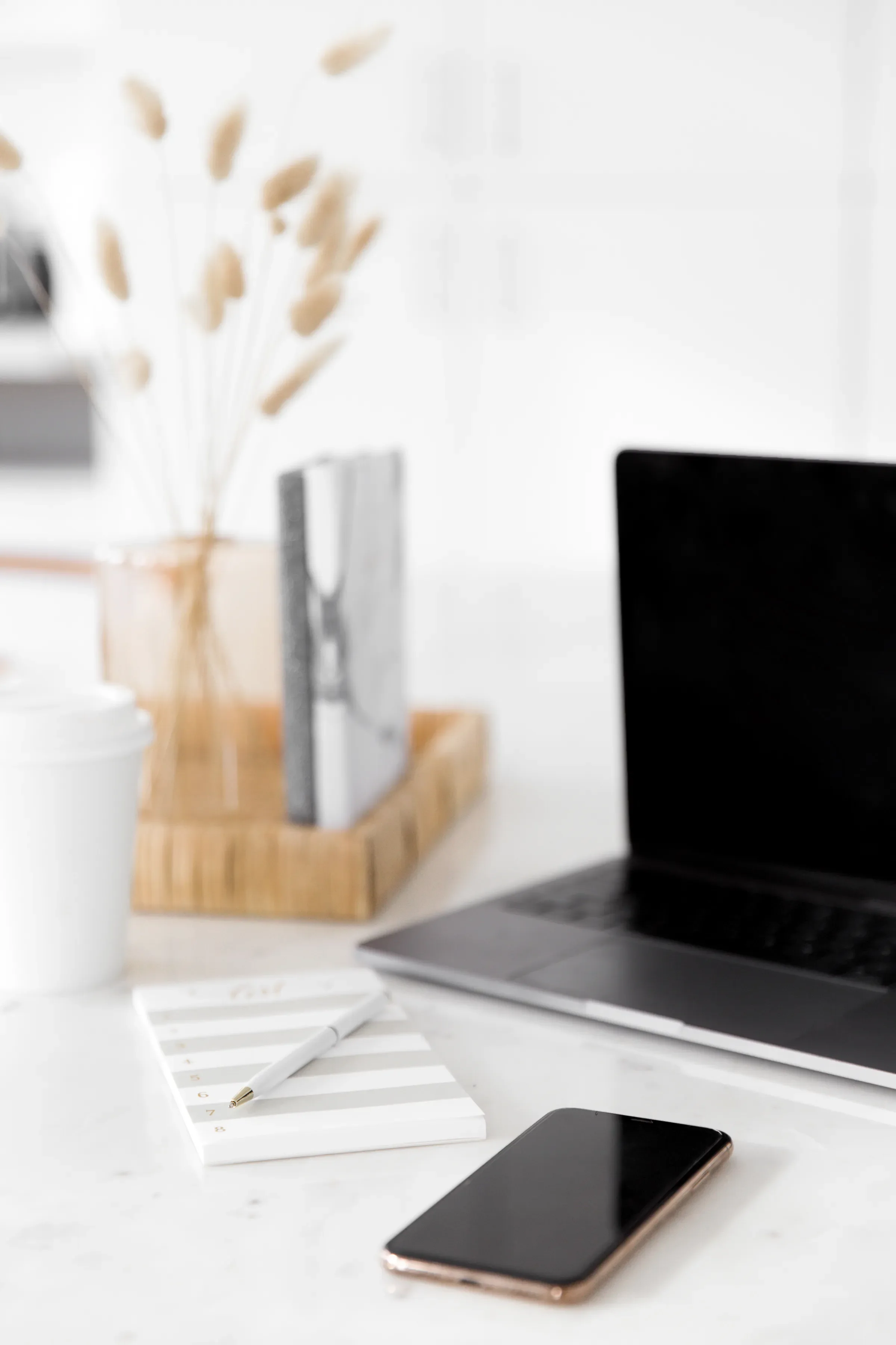 A cluttered white desk with a black smartphone, a white pen, a small notepad, a silver laptop, and a wooden tray holding a closed notebook, a white coffee cup, and a transparent vase with beige dried flowers, in a bright room.