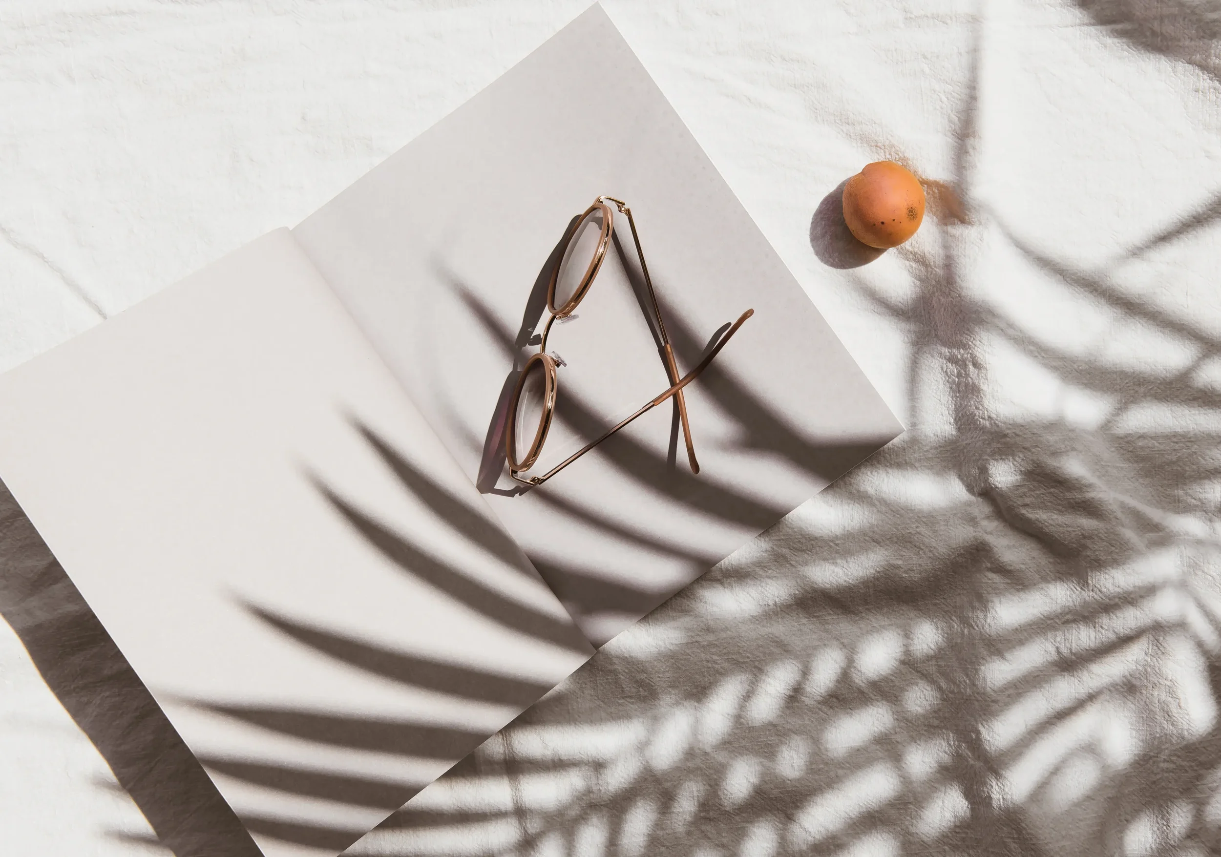 A pair of round sunglasses with brown frames resting on white paper, casting shadows on a textured white cloth surface, with a small potato nearby.