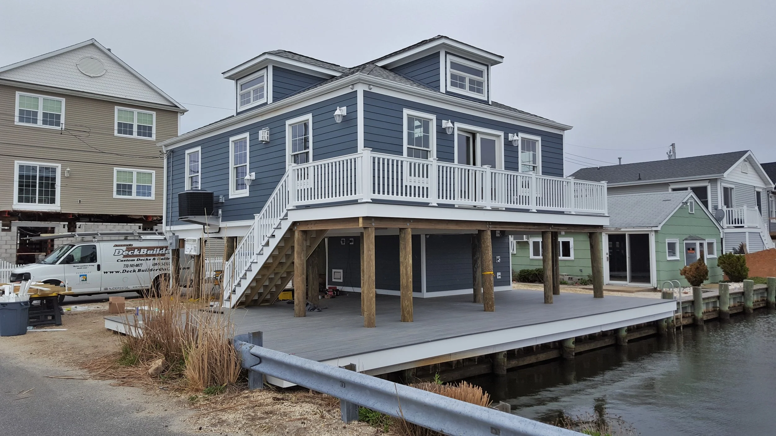 A house on stilts with a patio over water, located near other houses, under gray sky, with construction vehicles and tools present.