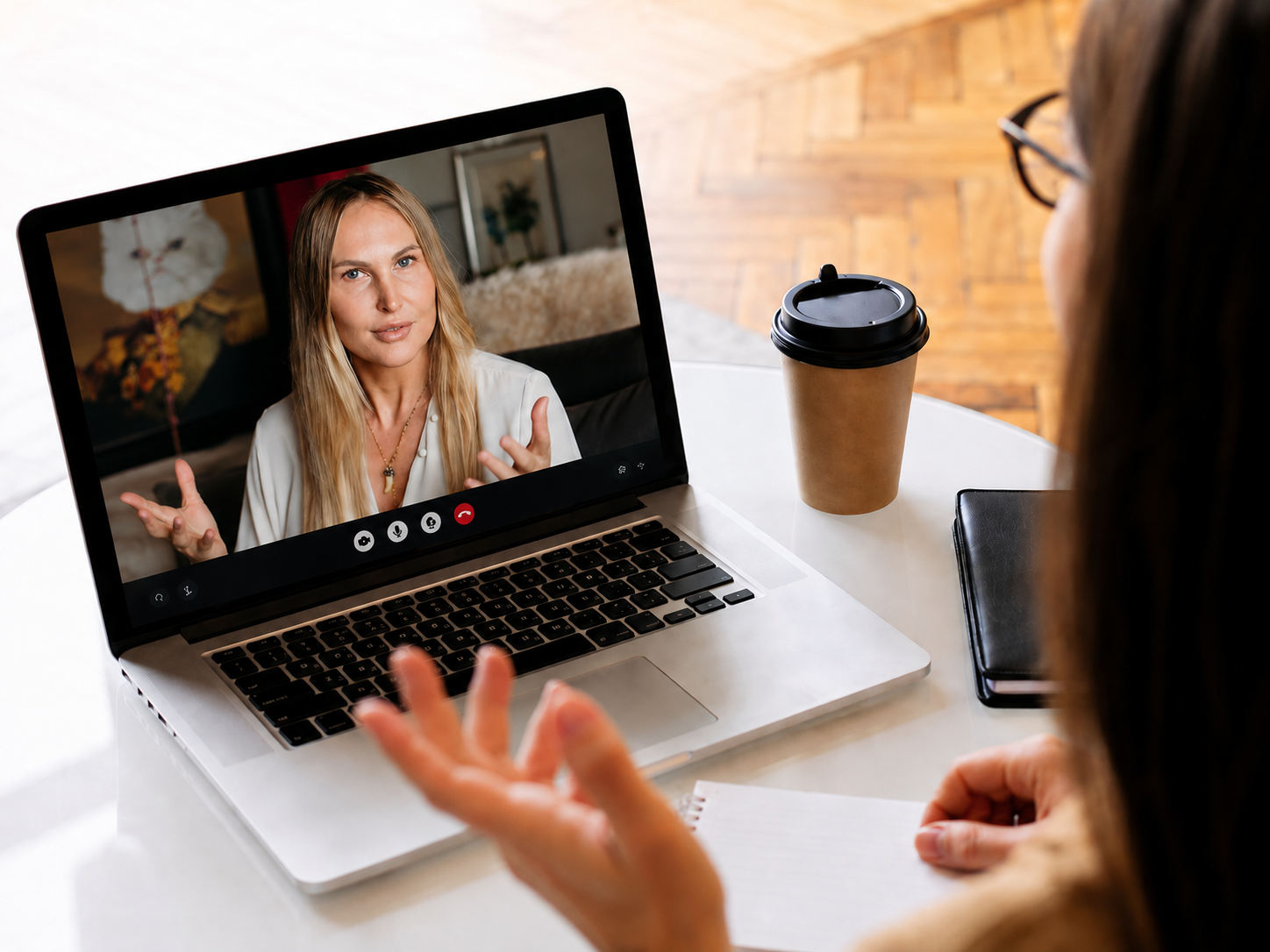 A woman is having a video call on her laptop with a blonde woman seen on the screen. The woman on the call is gesturing with her hands and talking. The setup includes a disposable coffee cup and a closed notebook on the table.