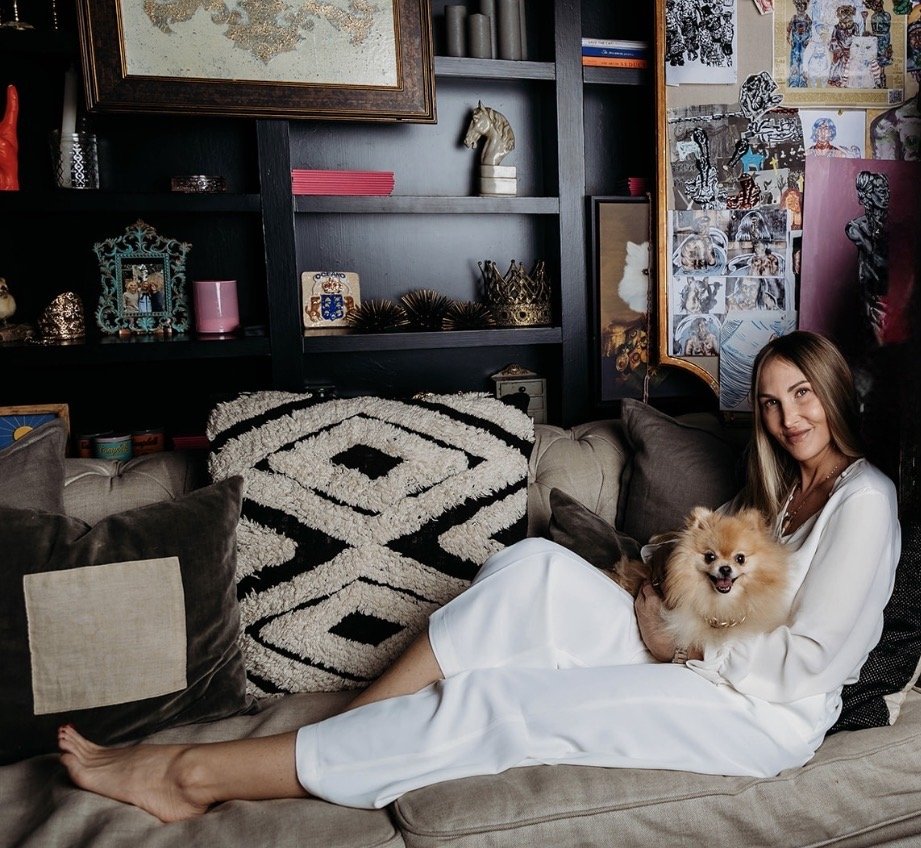 A woman in a white outfit sitting on a beige sofa, holding a fluffy Pomeranian dog, with a patterned blanket and pillows around her, in a room decorated with eclectic art and decor.