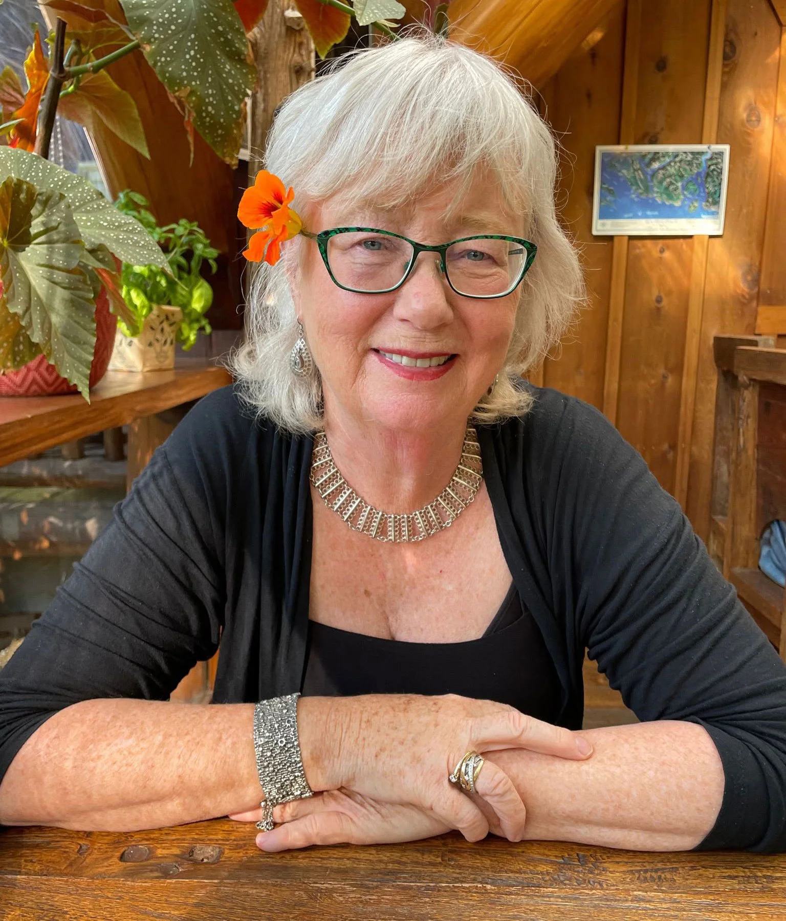 Author, Ann Svendsen wears a bright orange flower behind her ear, smiling at the camera sitting at a wooden table in a cozy, wood-paneled room with plants and a framed painting in the background.