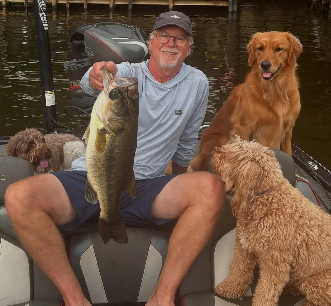 Man sitting on a boat holding a large fish, with three dogs around him and water in the background.