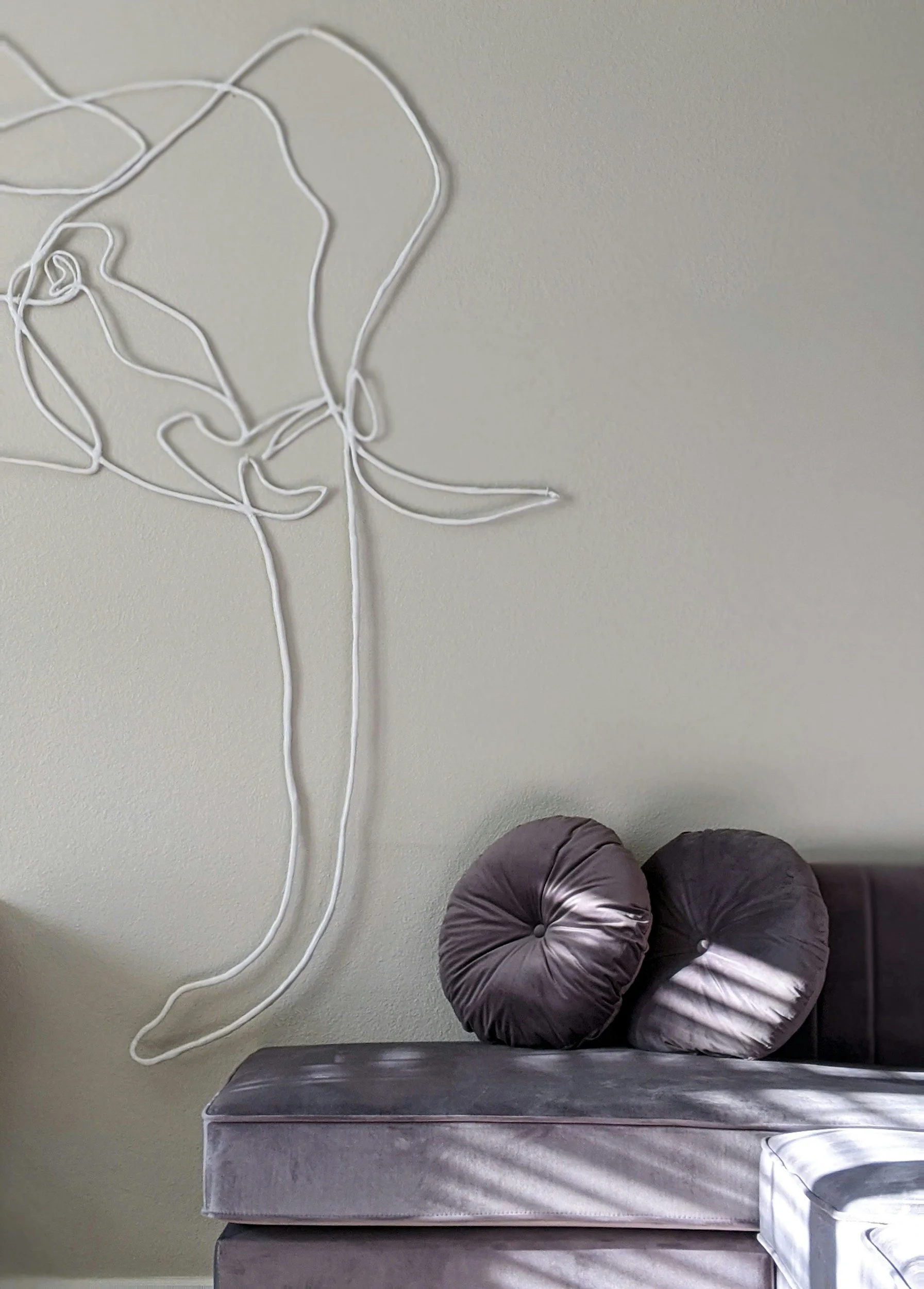 Blank beige wall with a wire art outline of an elephant trunk. A light gray couch with purple pillows in front.