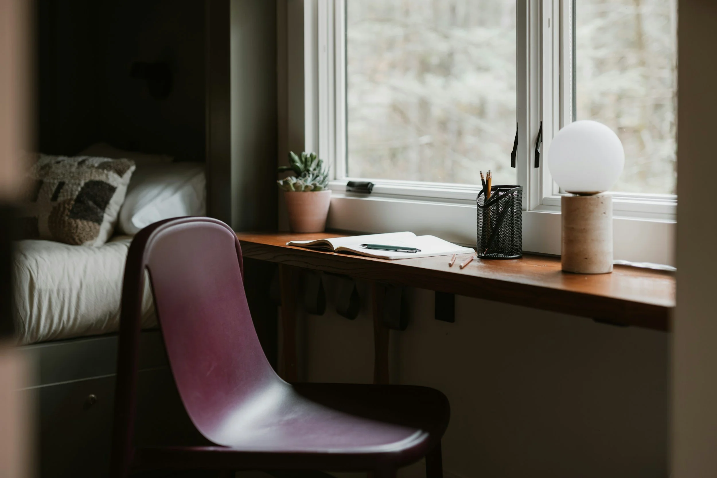 A desk near a window with a potted plant, a desk lamp, notebooks, and a pen, with a sofa and a pillow visible in the background.