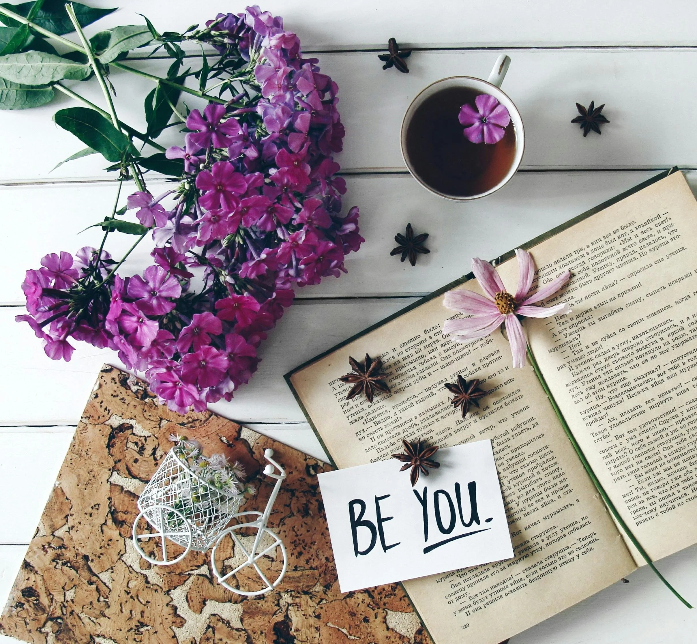 A flat lay of purple and pink flowers, star anise, a cup of tea with a purple flower, a botanical drawing, and a page from a book on a white wooden surface.