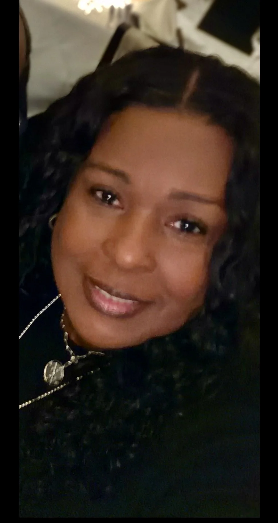 Close-up photo of a smiling woman with dark curly hair, wearing a black top and silver jewelry.