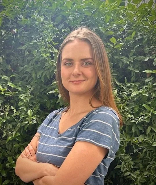 A woman with long, light brown hair and blue eyes standing outdoors in front of green bushes, wearing a blue and white striped t-shirt, smiling slightly.