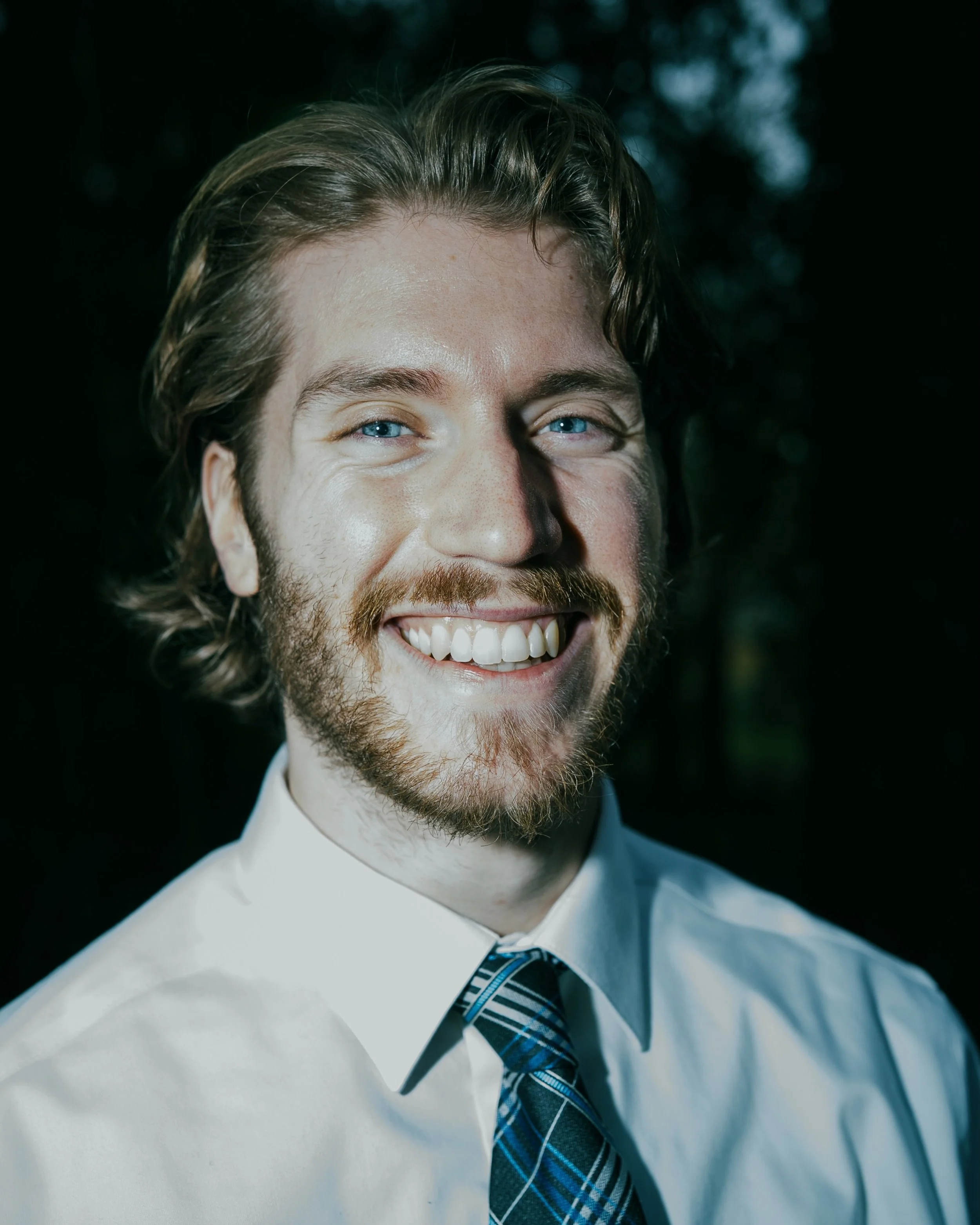 Close-up of a smiling man wearing a white shirt and a blue plaid tie, with a blurred dark background.