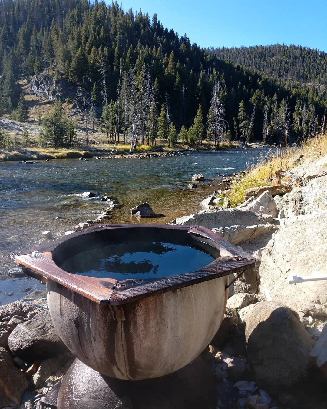 A hot tub filled with water on a rocky riverbank with a forested mountain in the background.
