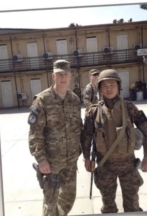 Two soldiers standing outside a military building, one in camouflage uniform with a patch on the sleeve and the other in tactical gear with a helmet, during daytime.