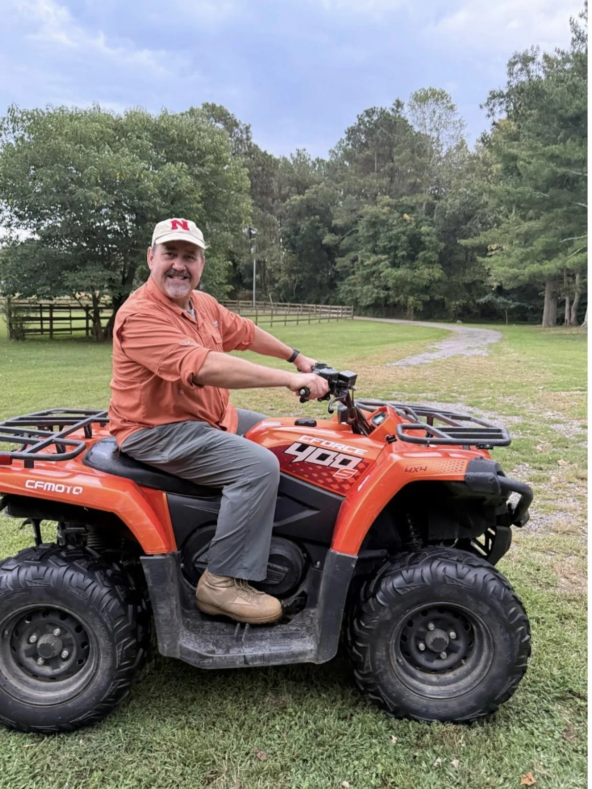 A man sitting on an orange all-terrain vehicle in a grassy outdoor area with trees and a dirt path in the background. The man is wearing a beige cap with a red 'N', an orange shirt, gray pants, and tan boots, and he's smiling at the camera.