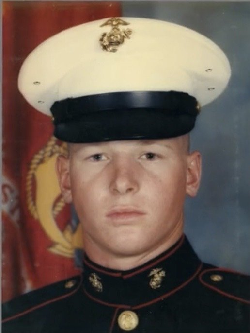 A young man in a Marine Corps uniform wearing a white drill instructor hat with a gold emblem, standing in front of a red and yellow military flag.