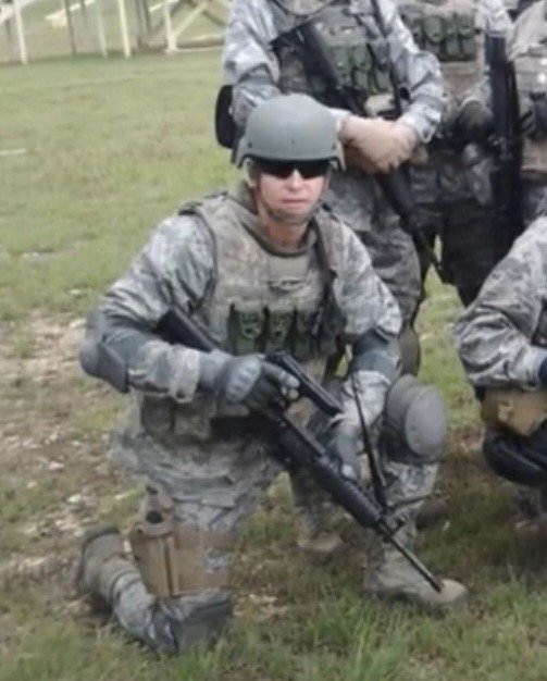 A soldier in camouflage uniform, kneeling on the grass, holding a rifle, wearing a helmet and sunglasses.