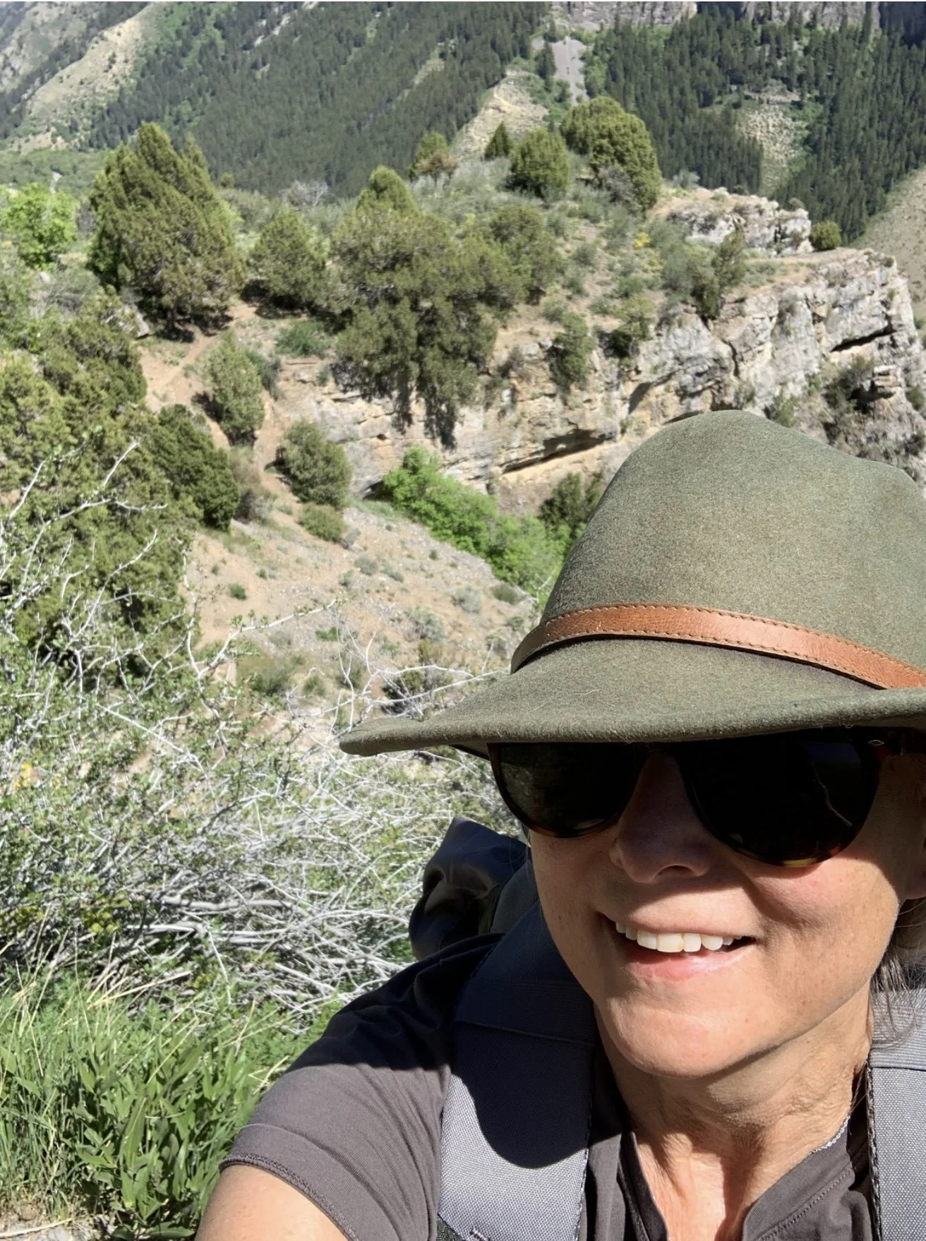 A person wearing a hat and sunglasses smiling, with a mountain and trees in the background.