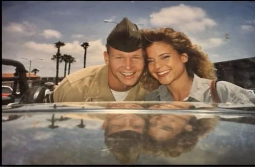 A man in military uniform and a woman with curly hair smiling and leaning over a car windshield, with their reflections visible on the glass, in a sunny outdoor parking lot.