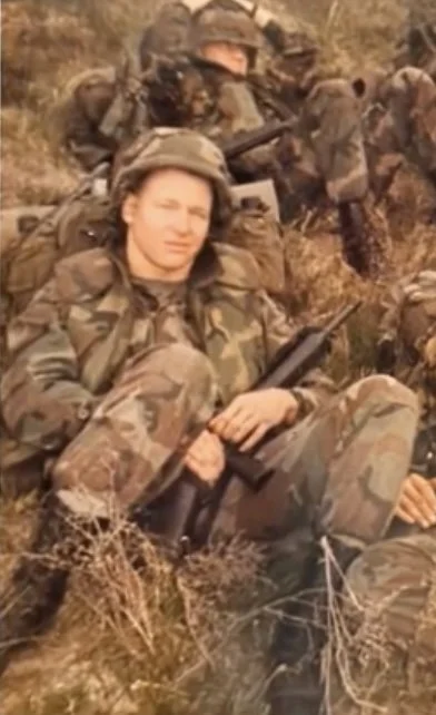A group of soldiers in camouflage uniforms resting outdoors on a grassy hillside. One soldier, a woman, is sitting in the foreground holding a rifle, with others seated behind her.
