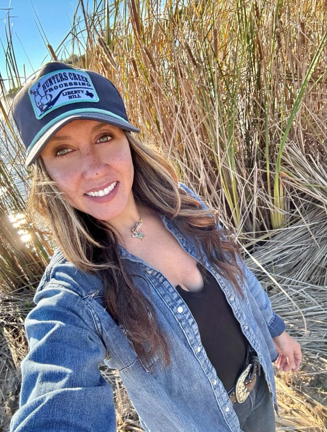 Woman wearing a trucker hat with the text 'Hunters Creek Progressing Liberty Hill' and a deer logo, smiling in a field of tall, dried grass on a sunny day.