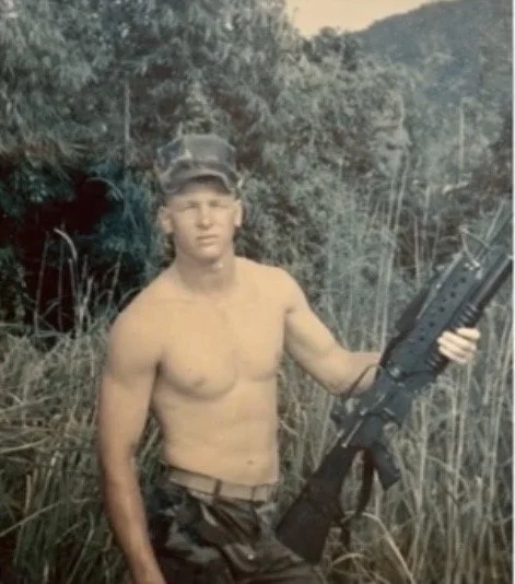 A shirtless young man wearing a camouflage cap and sitting outdoors in tall grass, holding a firearm in his right hand.