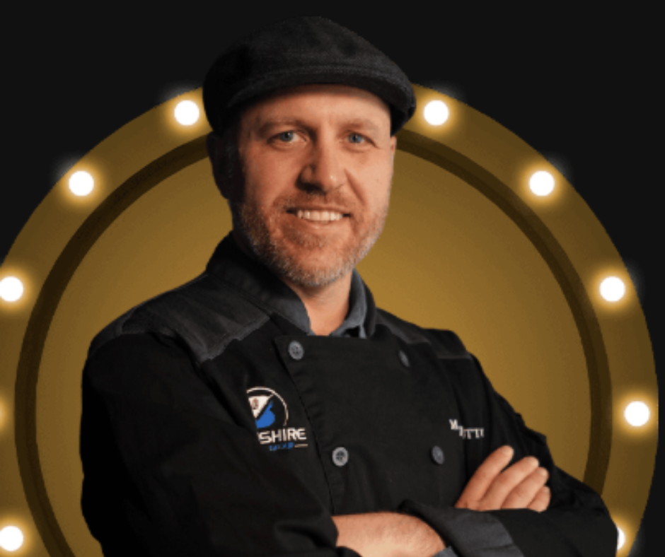 A man in a black chef's coat and a flat cap stands with arms crossed in front of a lit marquee circle backdrop.