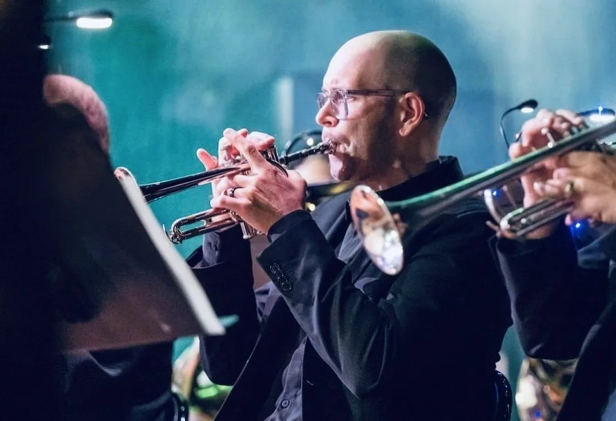 A man playing the trumpet in an orchestra, wearing glasses and a black suit.