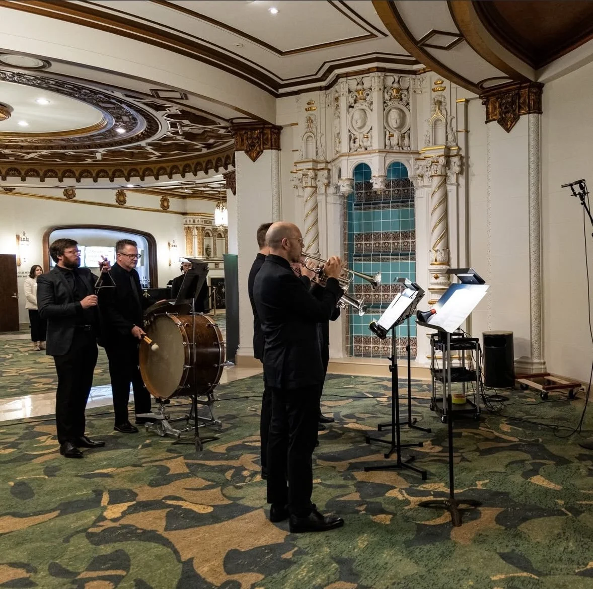 A small ensemble performing indoors, including trumpet and percussion instruments, in a room with ornate architecture and a decorative ceiling.