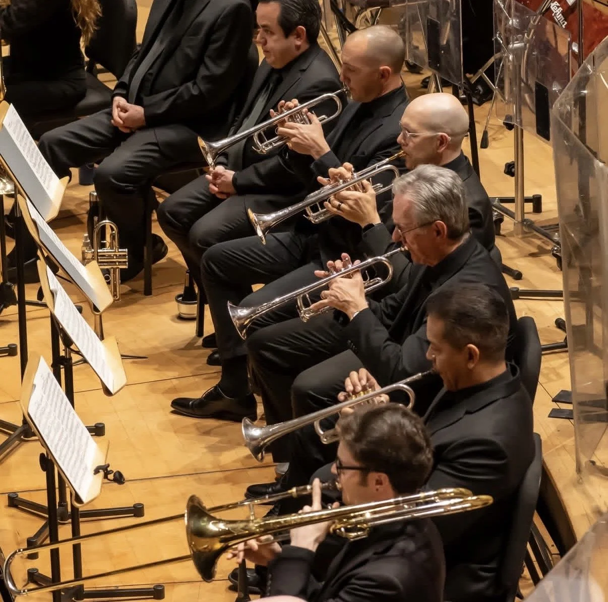 Orchestra musicians seated in a concert hall playing trumpets during a performance.
