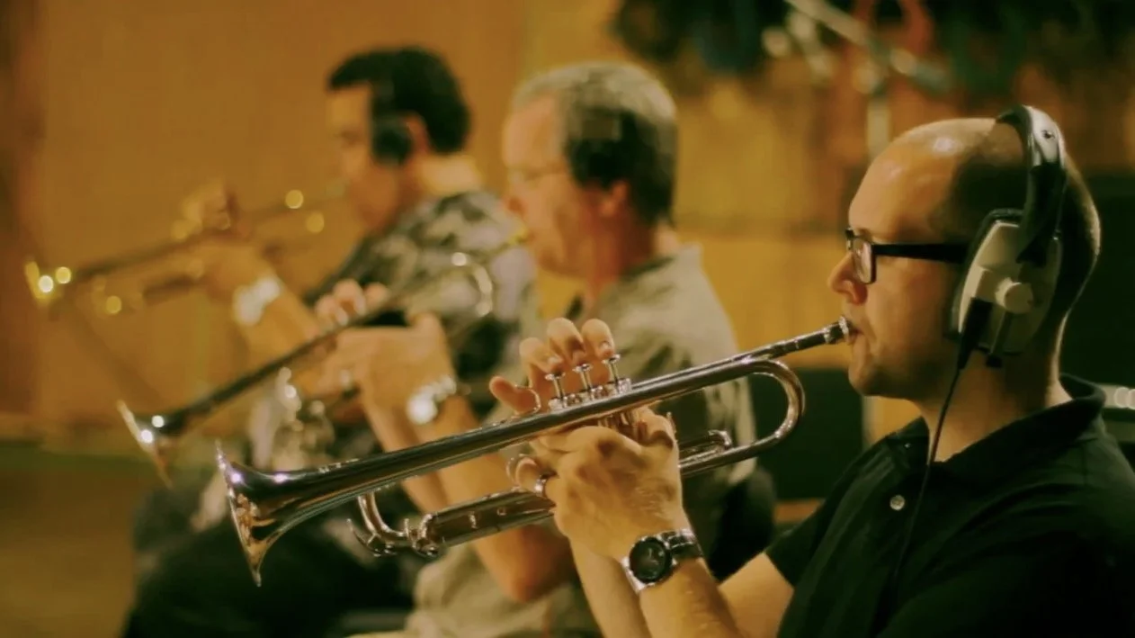 Three male musicians in a recording studio, playing trumpets while wearing headphones, with warm lighting in the background.