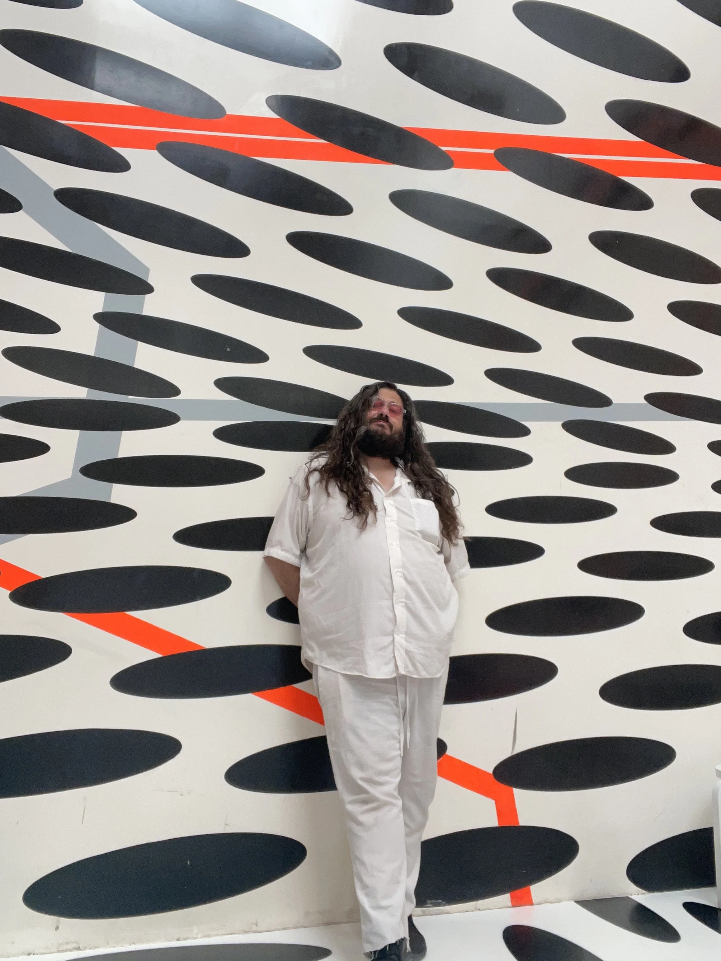 A man with long curly hair, beard, glasses, white shirt, and white pants standing against an abstract black oval pattern background with orange and gray lines.
