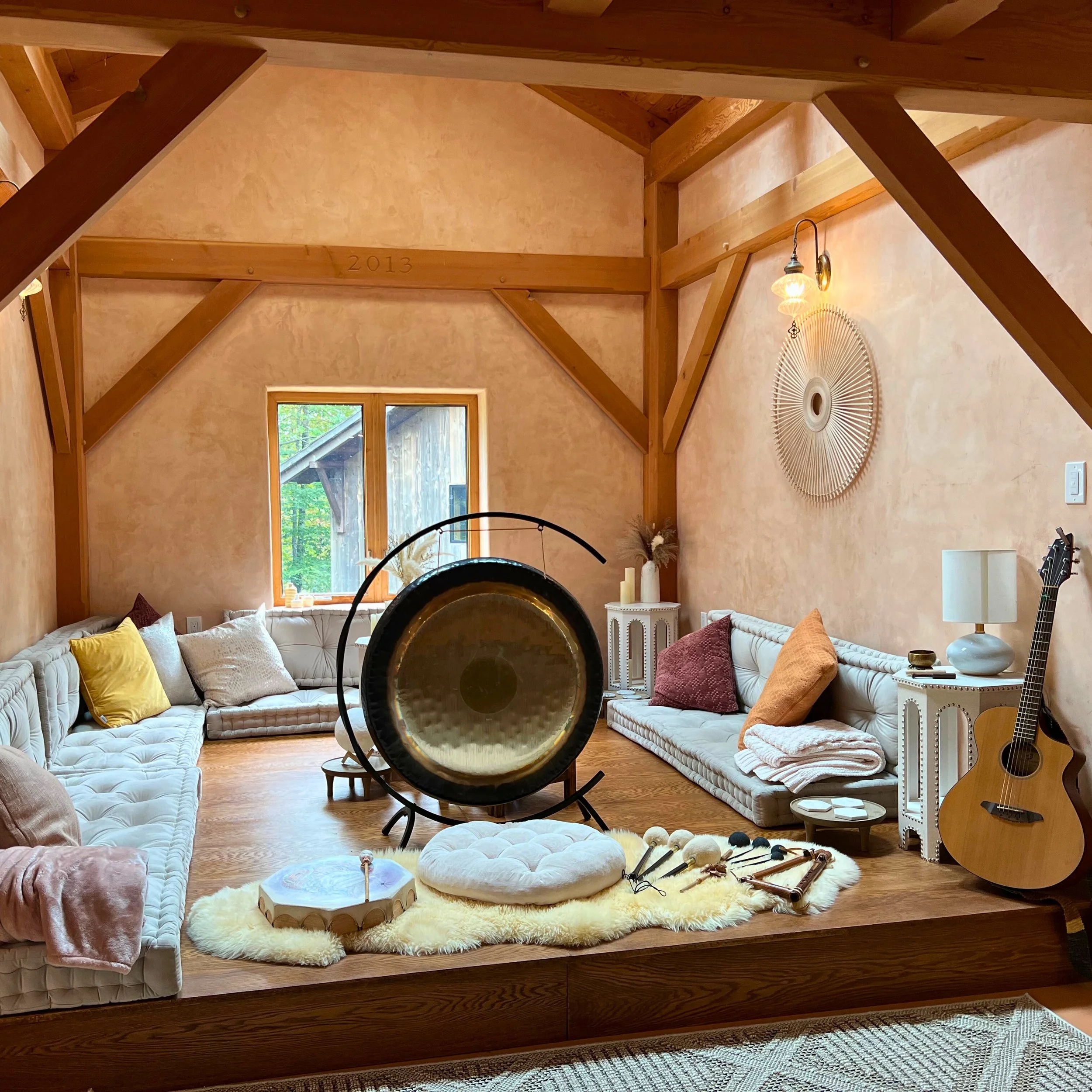 A cozy living room with wooden beams, a large window, a white sectional sofa with yellow and maroon pillows, a fluffy round rug, musical instruments, and wall decorations.