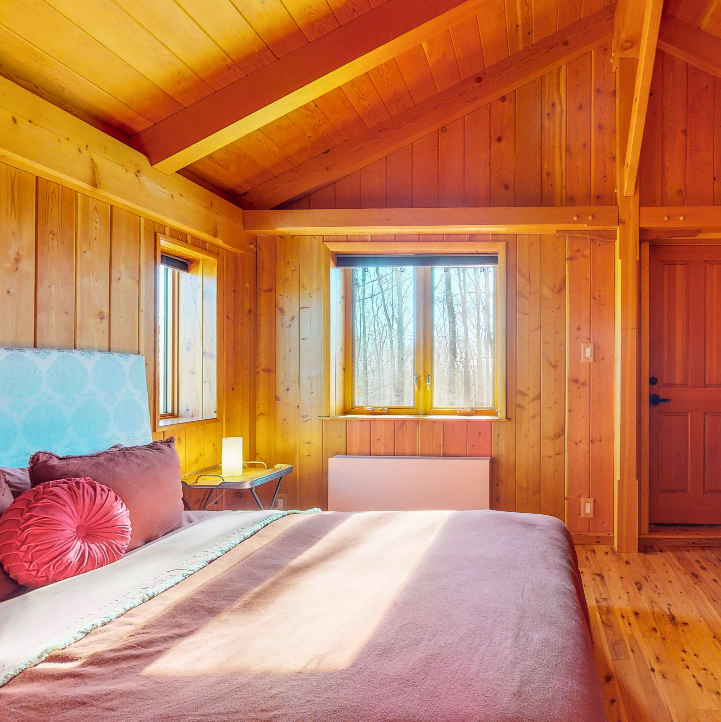 A bedroom with wood-paneled walls and ceiling, a window showing trees outside, a bed with pink pillows, and a nightstand with a lamp.